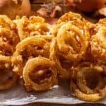 Golden, crispy onion rings stacked on parchment paper, with whole onions placed in the background.