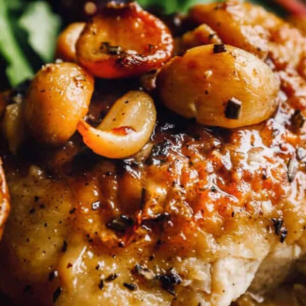 Close-up of a savory dish featuring roasted chicken with a glossy glaze, topped with caramelized onions and served alongside crispy potato slices. Fresh greens peek in the background.