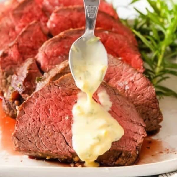 Slices of medium-rare roast beef are arranged on a white plate. A spoon drizzles a creamy sauce over the meat. Fresh rosemary garnishes the dish, and a striped cloth is visible to the side.