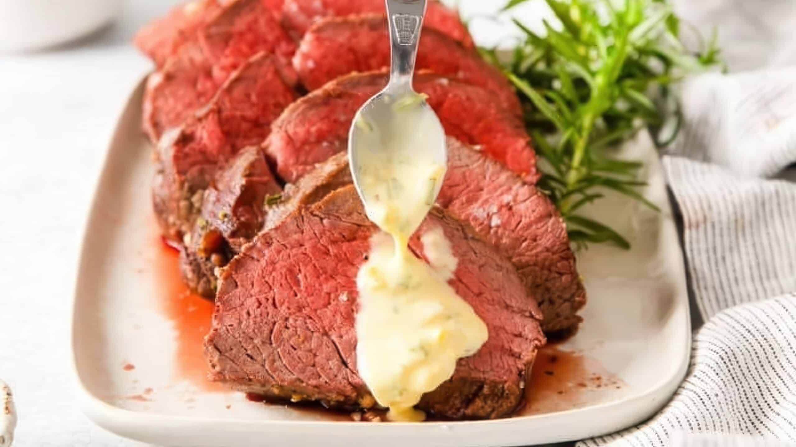 Slices of medium-rare roast beef are arranged on a white plate. A spoon drizzles a creamy sauce over the meat. Fresh rosemary garnishes the dish, and a striped cloth is visible to the side.