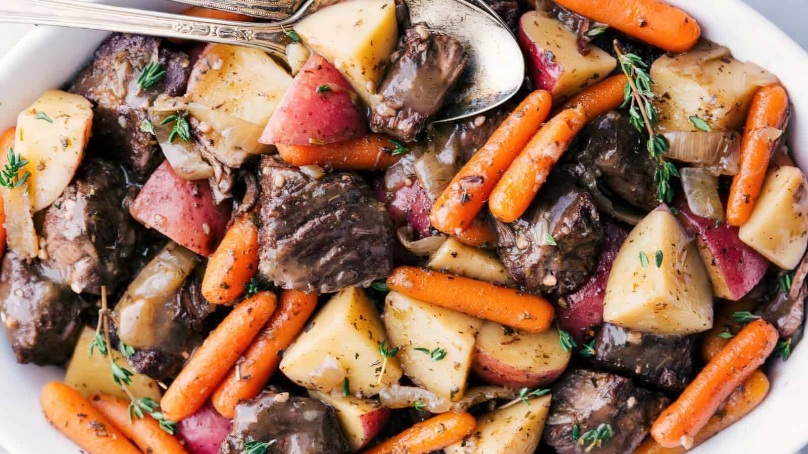A hearty dish featuring chunks of tender beef, roasted red potatoes, and glazed baby carrots, garnished with fresh thyme sprigs. The meal is served in a white bowl with two silver serving spoons resting on top.