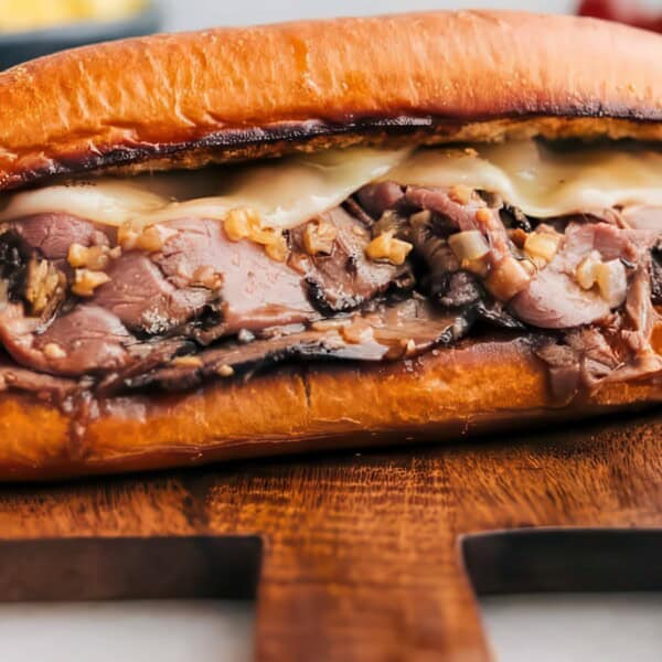 Close-up of a juicy roast beef sandwich with melted cheese on a toasted bun, placed on a wooden board. The sandwich is filled with thinly sliced beef and topped with onions. In the blurred background, there are grapes and a bowl, adding a touch of color.