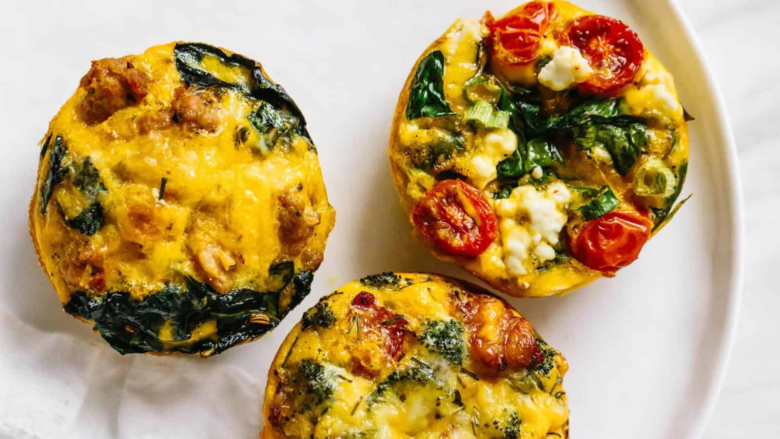 Three savory egg muffins on a white plate, topped with spinach, tomatoes, and crumbled cheese. The muffins are golden brown and studded with greens and vegetables, creating a colorful and appetizing presentation.