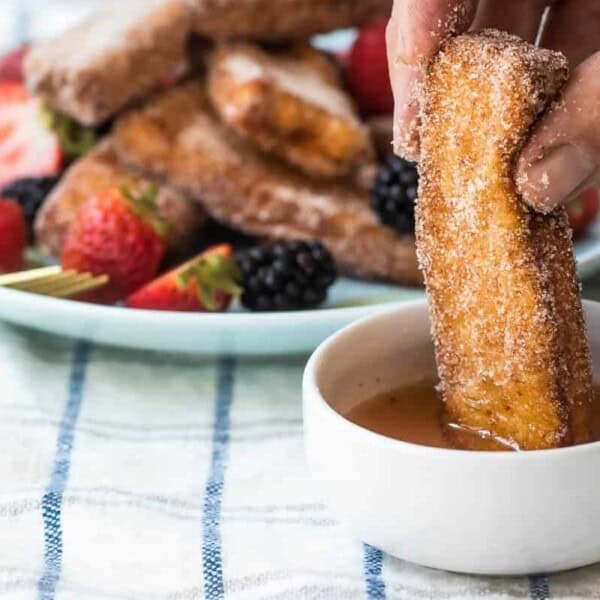 A person dipping a piece of cinnamon sugar-coated French toast stick into a small bowl of syrup. In the background, there's a plate of more French toast sticks, garnished with sliced strawberries and blackberries on a striped cloth.