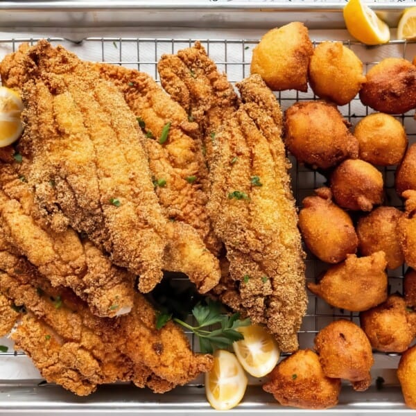 A tray of golden fried catfish fillets and hushpuppies garnished with lemon wedges, accompanied by a small bowl of coleslaw in the corner. The food is arranged on a wire rack over a baking sheet, with a striped napkin nearby.