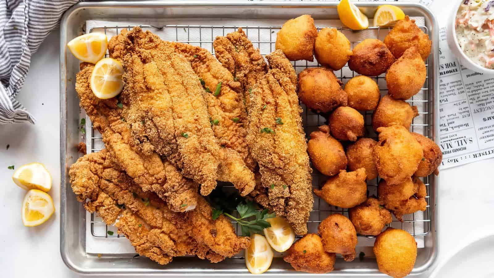 A tray of golden fried catfish fillets and hushpuppies garnished with lemon wedges, accompanied by a small bowl of coleslaw in the corner. The food is arranged on a wire rack over a baking sheet, with a striped napkin nearby.