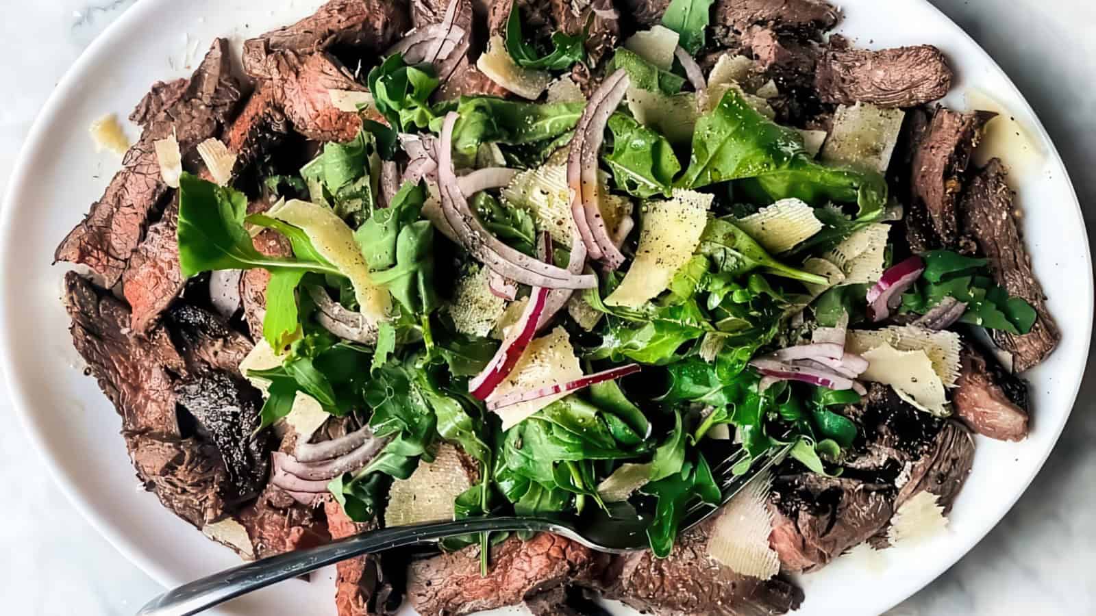 A plate of sliced steak topped with a mixed green salad, featuring arugula, red onion slices, and parmesan shavings, with a fork resting on the plate. Served with