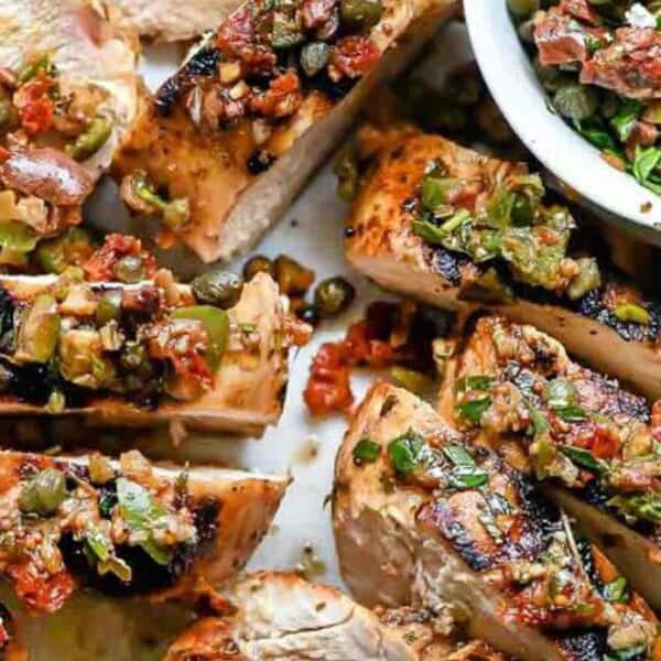 Grilled chicken breast slices topped with a mixture of chopped olives, capers, and herbs. A small bowl with more olive mixture is on the side, along with lemon wedges. The dish is garnished with crumbled cheese.