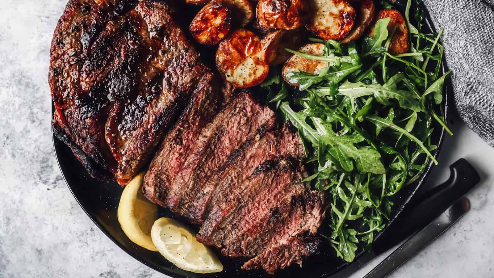 A plate with sliced grilled steak, roasted potatoes, and fresh arugula, garnished with lemon wedges. A fork and knife are placed beside the plate on a textured gray surface.