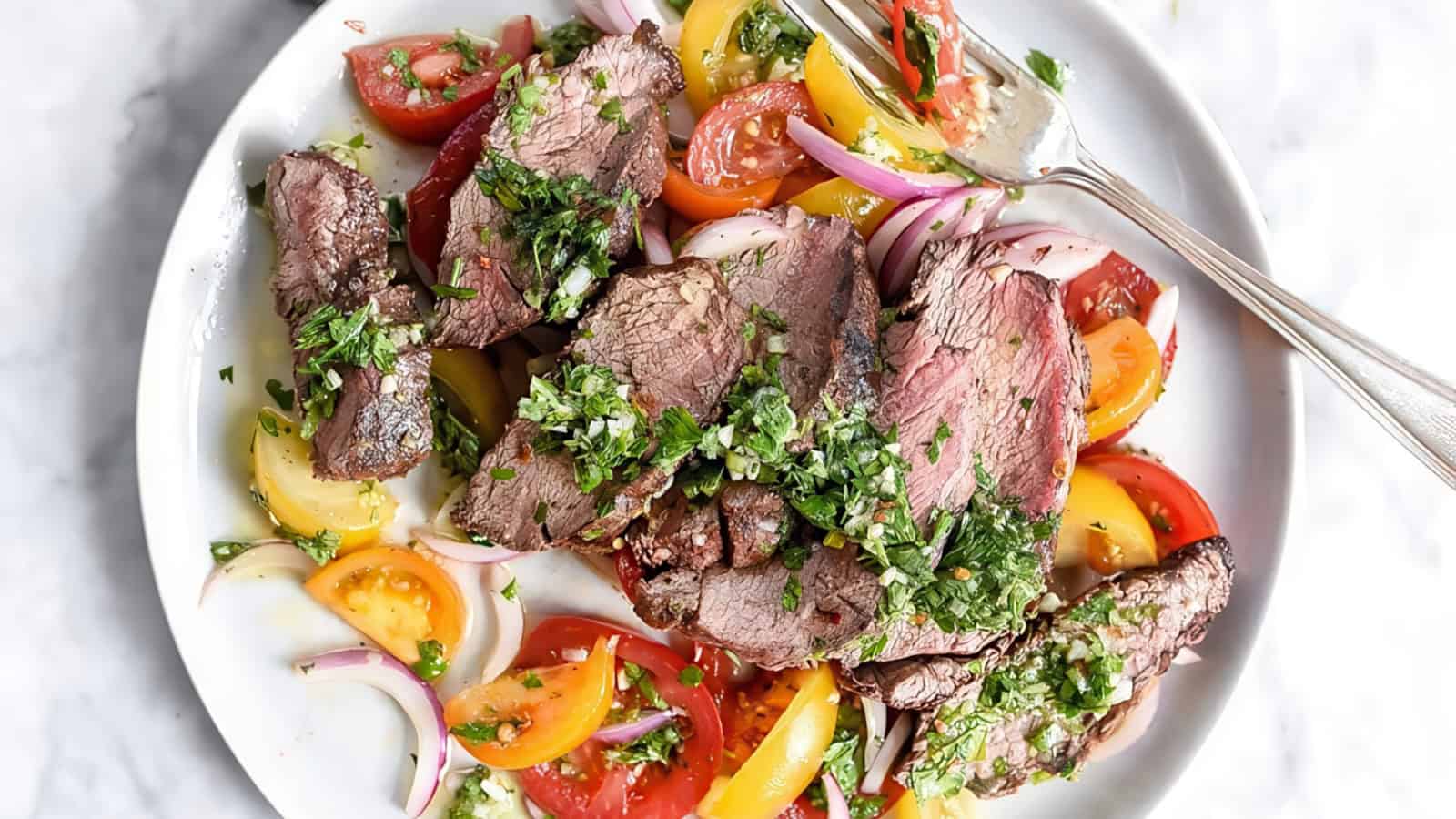 A plate of sliced grilled steak topped with chopped herbs. It's served over a colorful salad of yellow and red cherry tomatoes, red onion slices, and crumbled feta cheese. Two forks rest on the side of the plate.