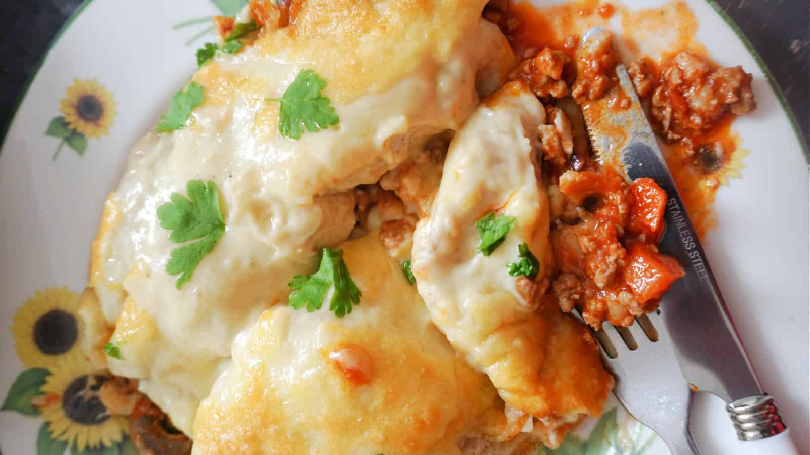 A plate of cheesy lasagna topped with melted cheese and garnished with fresh parsley. A fork and knife rest on the side, with a bit of tomato sauce and cheese on the silverware. The plate has a sunflower pattern.