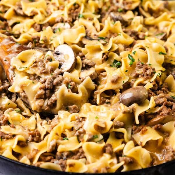 A skillet filled with beef stroganoff, featuring wide egg noodles, ground beef, and mushrooms in a creamy sauce. A wooden spoon rests in the skillet, garnished with chopped parsley.