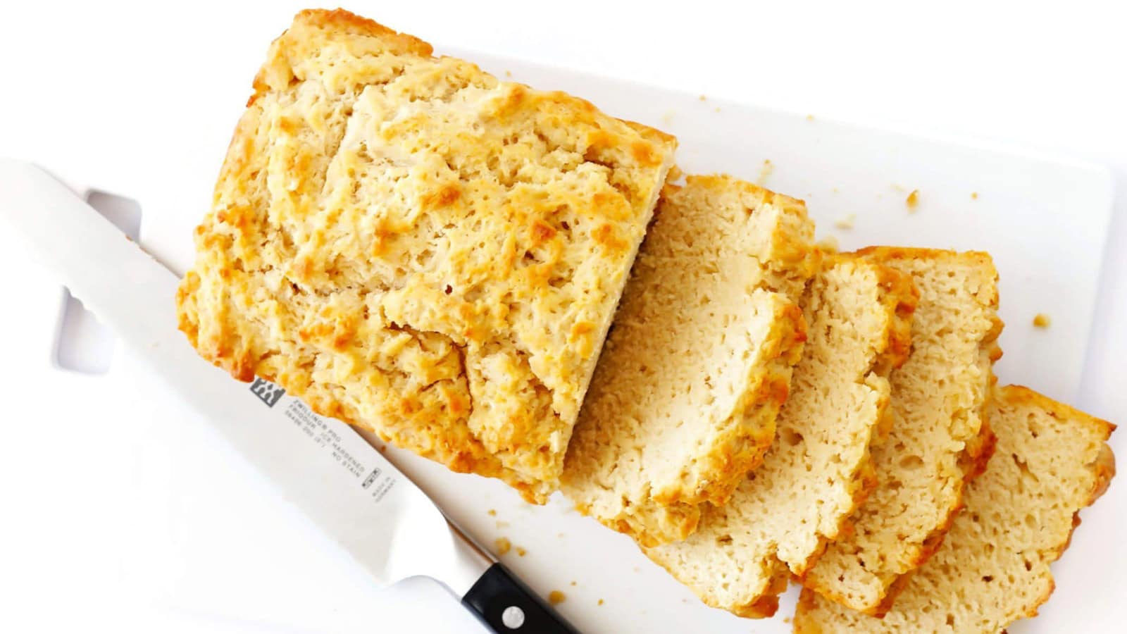 A loaf of freshly baked bread is partially sliced on a white cutting board. A bread knife with a black handle rests beside the loaf. The bread has a golden crust and a soft, fluffy interior.