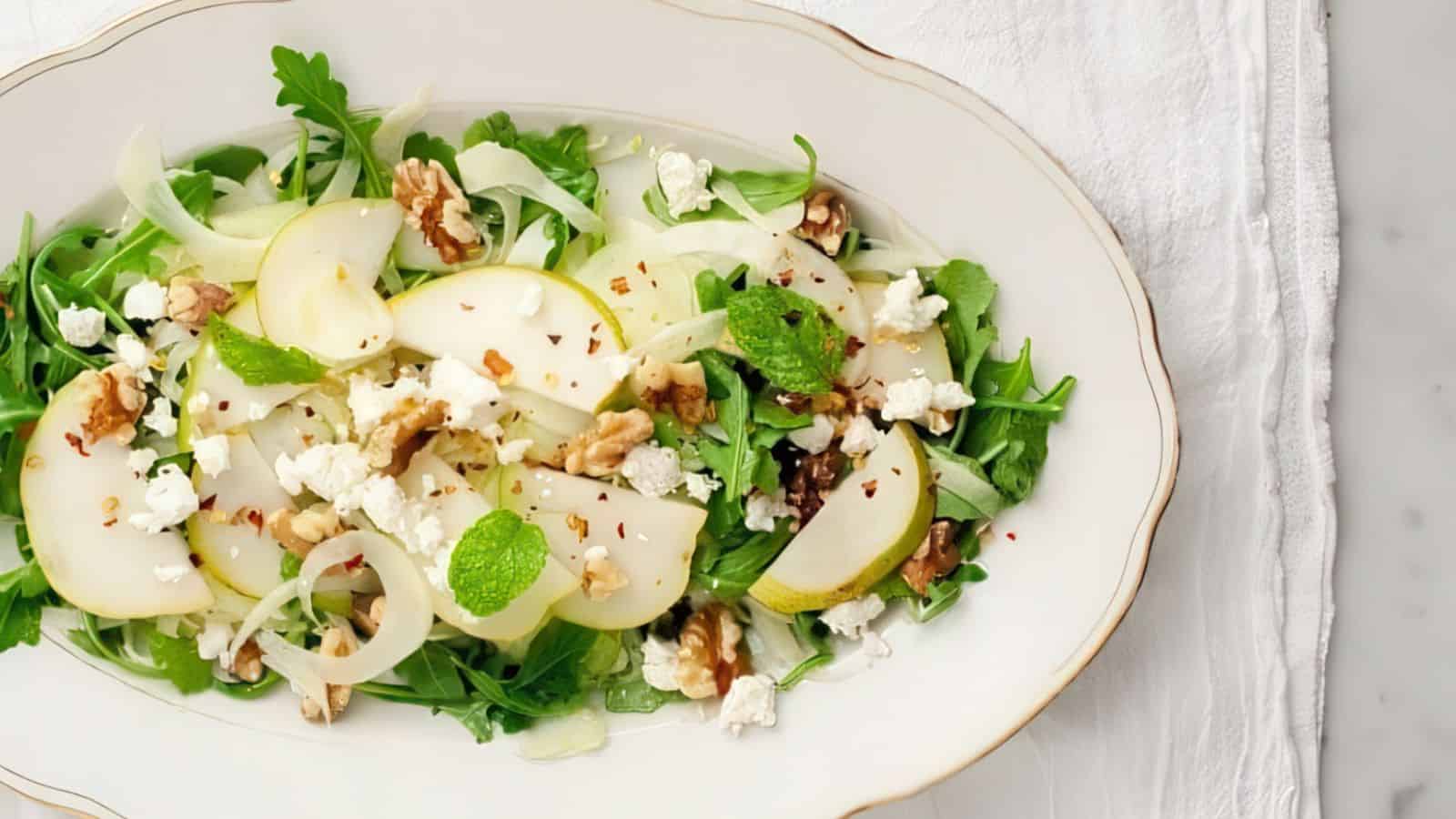 A white oval plate with a salad of arugula, pear slices, walnuts, feta cheese, and mint leaves on a light cloth background. The salad is garnished with a sprinkle of red pepper flakes and drizzled with a light dressing.