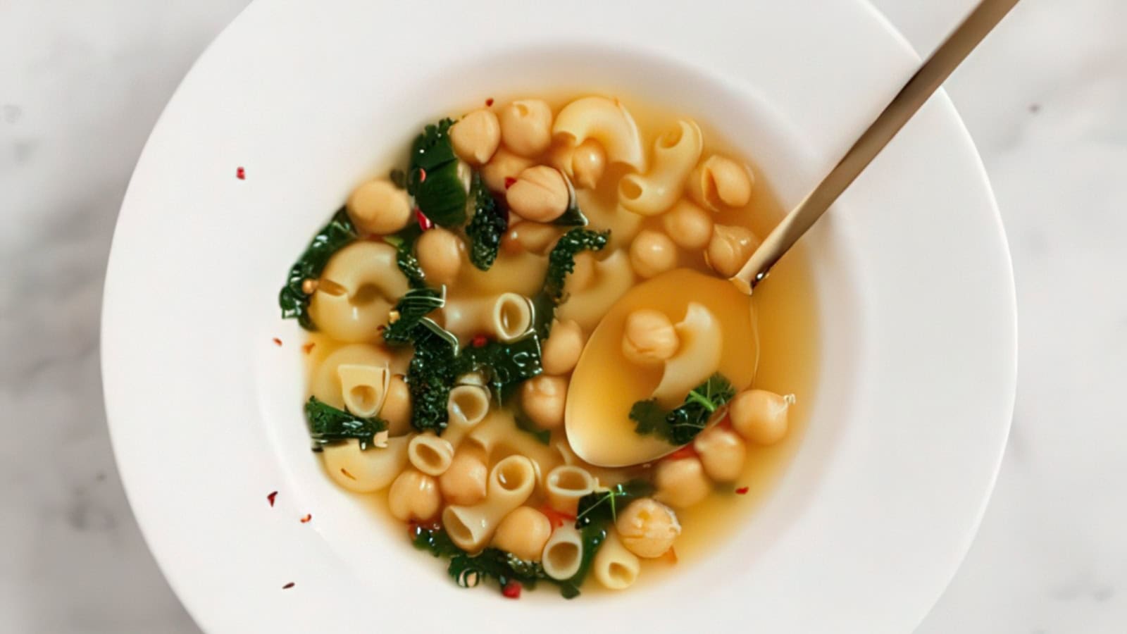 A white bowl of soup containing chickpeas, small shell pasta, and dark green kale in a clear broth. A spoon is lifting a few chickpeas from the soup. Sprinkles of red pepper flakes are visible on the surface.