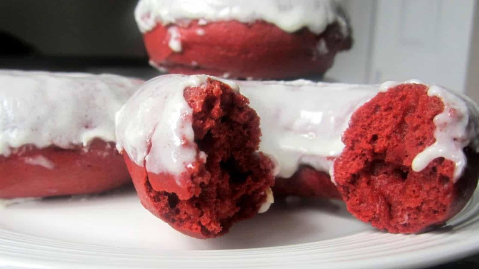 A close-up of red velvet donuts with white icing on a white plate. One donut is bitten, revealing its soft, red interior. The icing glistens under the light, creating a tempting, indulgent treat.
