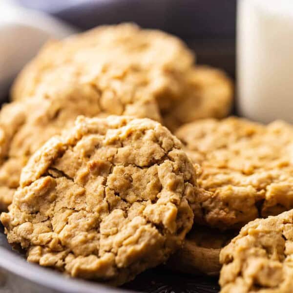 A plate of oatmeal cookies is stacked closely together. The cookies have a rough, textured surface. In the background, a glass of milk is partially visible, adding a cozy, homemade feel to the setting.