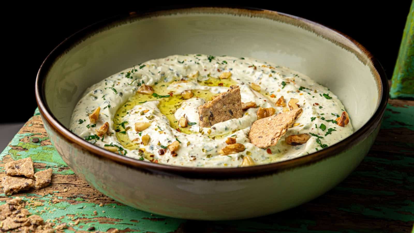 A bowl of creamy dip topped with olive oil, chopped nuts, and herbs, garnished with crispy cracker pieces, placed on a rustic wooden surface.