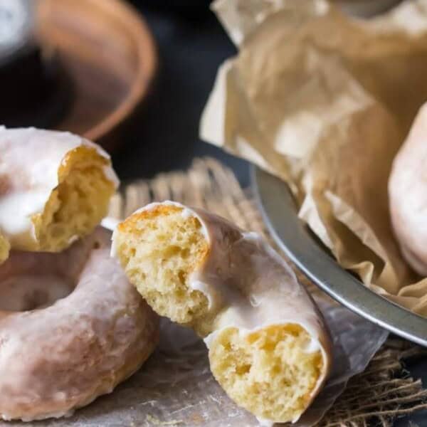 A close-up of several glazed doughnuts on parchment paper. One doughnut is broken in half, revealing its fluffy interior. A tin with more doughnuts is visible in the background, lined with parchment paper.