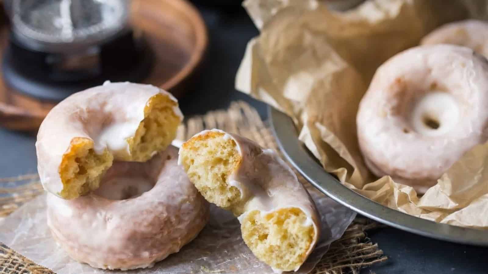 A close-up of several glazed doughnuts on parchment paper. One doughnut is broken in half, revealing its fluffy interior. A tin with more doughnuts is visible in the background, lined with parchment paper.