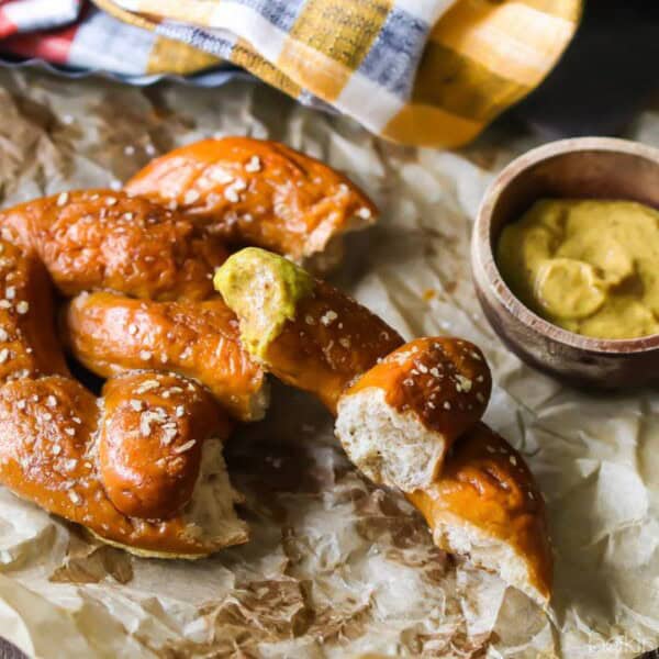A soft pretzel with coarse salt, partially broken on parchment paper, sits beside a small wooden bowl of mustard.