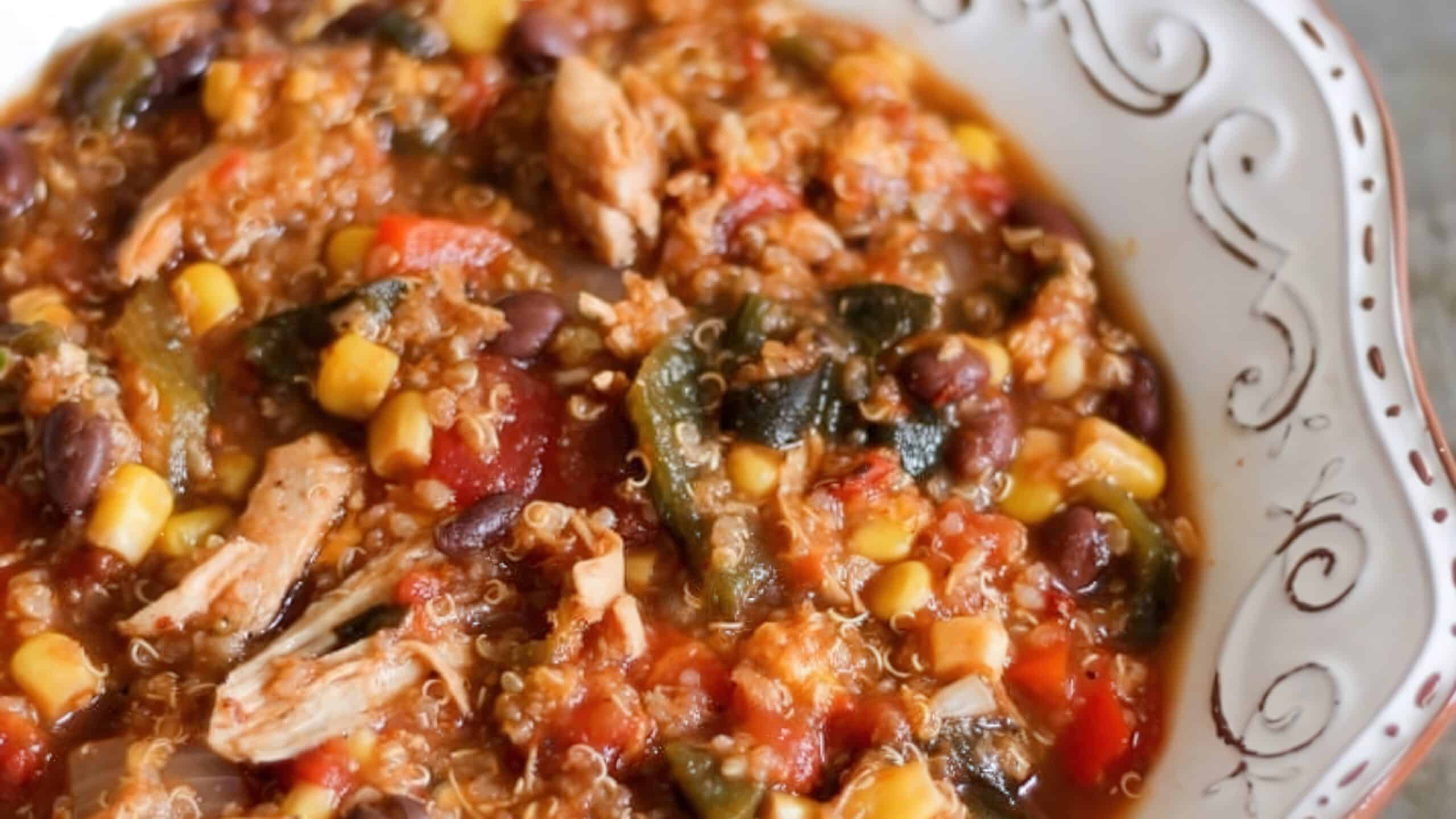 A bowl of vibrant quinoa chili with chunks of chicken, black beans, corn, tomatoes, and zucchini, all in a rich, savory broth, served in a decorative white bowl.