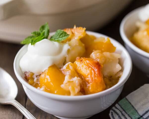 A small white bowl filled with peach cobbler topped with a dollop of whipped cream and garnished with a mint sprig. A spoon and a checkered napkin are placed nearby on a wooden table.