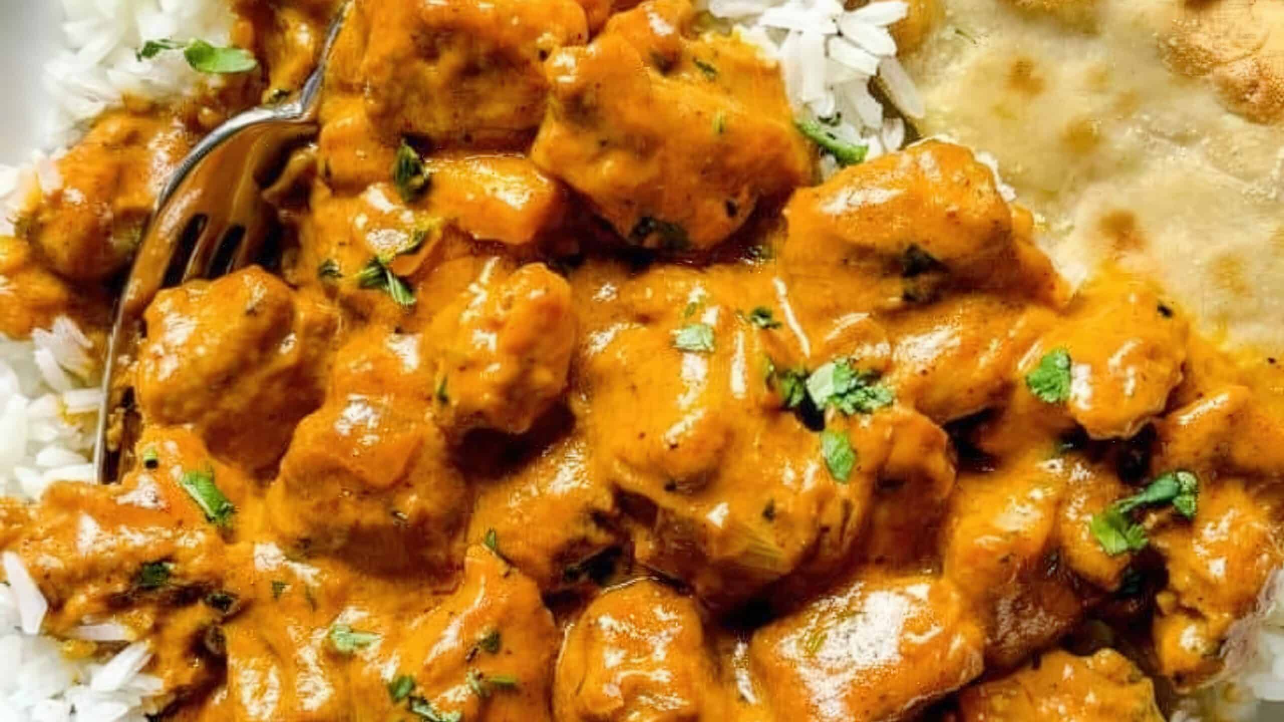 A close-up of a plate with creamy orange butter chicken garnished with herbs. The dish is served with white rice and a piece of naan bread, with a fork partially visible on the side.