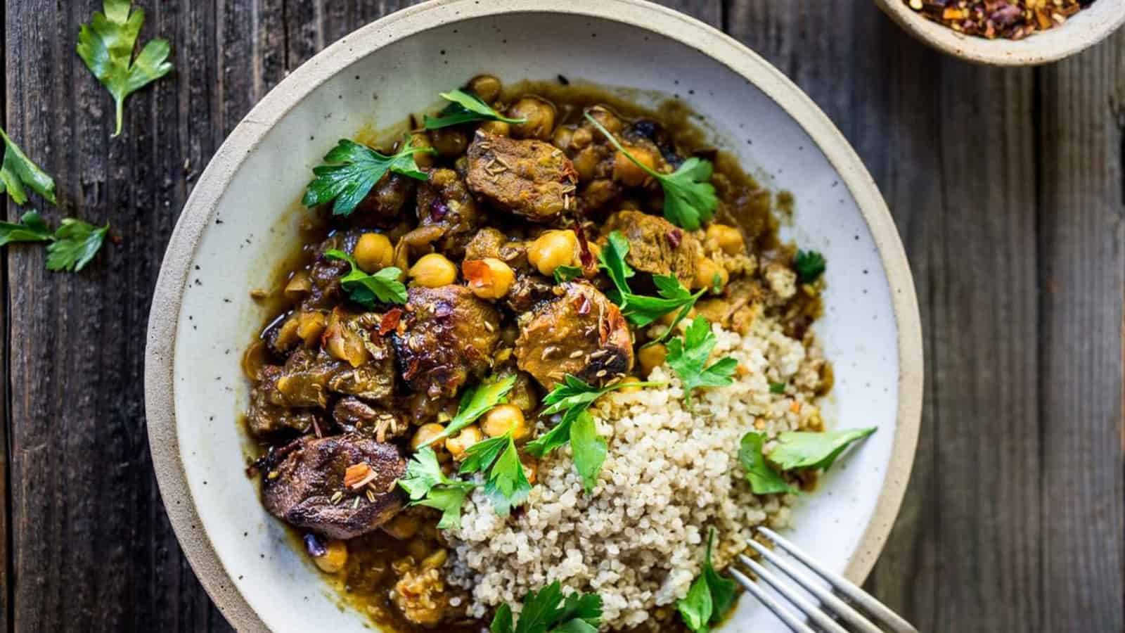 A bowl of beef and chickpea stew topped with fresh parsley, served with a side of quinoa. The dish is set on a rustic wooden table, with additional parsley leaves scattered around. A fork rests on the edge of the bowl.