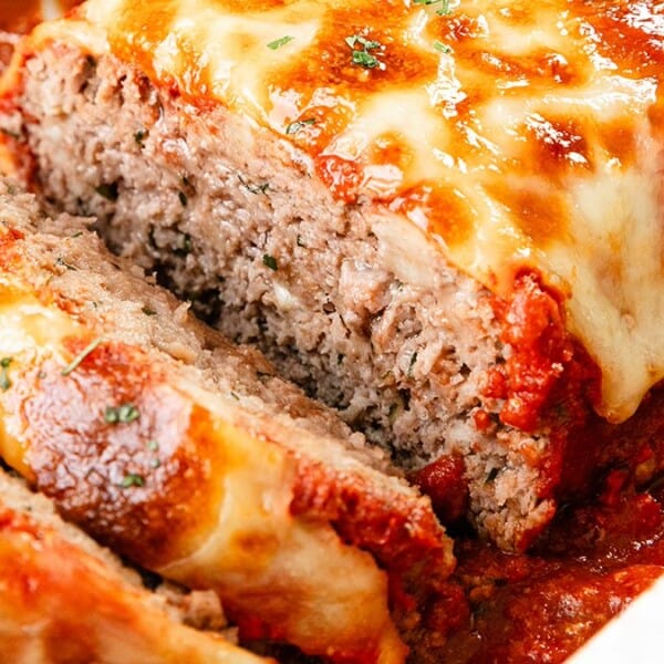 A close-up of a meatloaf topped with melted cheese and garnished with herbs. It is partially sliced, revealing a moist, textured interior. The dish is set in a pool of rich red sauce.