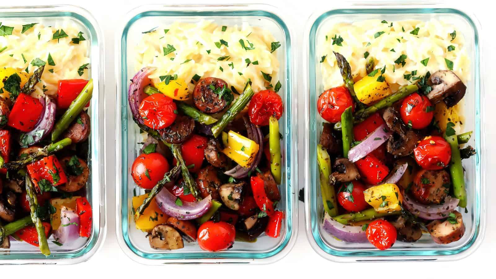 Three glass containers filled with a colorful arrangement of sliced sausages, cherry tomatoes, red and yellow bell peppers, red onion, asparagus, and orzo pasta, garnished with fresh herbs.