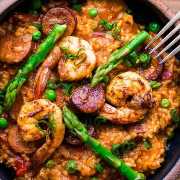 A bowl of risotto with shrimp, sausage, green peas, asparagus, and chopped herbs is eaten with a fork.
