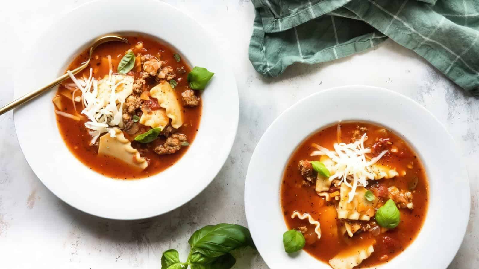 Two bowls of lasagna soup topped with shredded cheese and fresh basil on a light surface. A gold spoon rests in one bowl, and a green cloth is placed nearby.