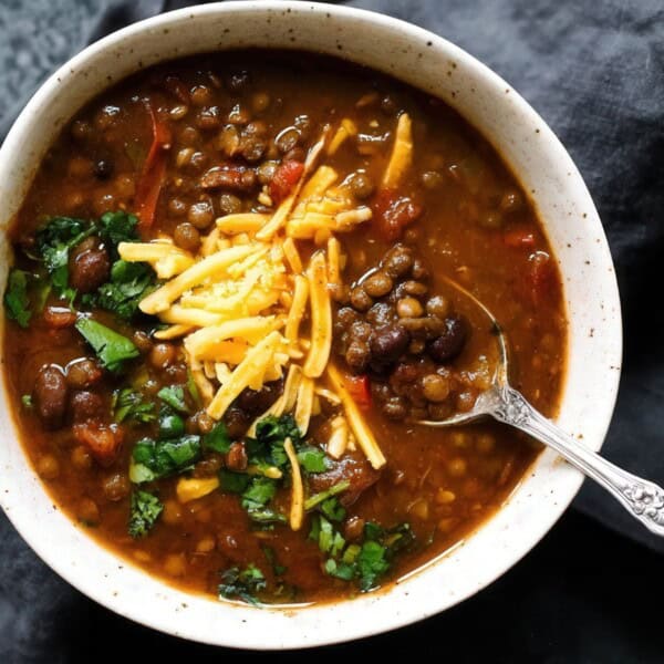 A bowl of lentil soup topped with shredded cheese and chopped herbs. A spoon is resting in the bowl. The soup is placed on a dark cloth, with part of another bowl visible in the corner.