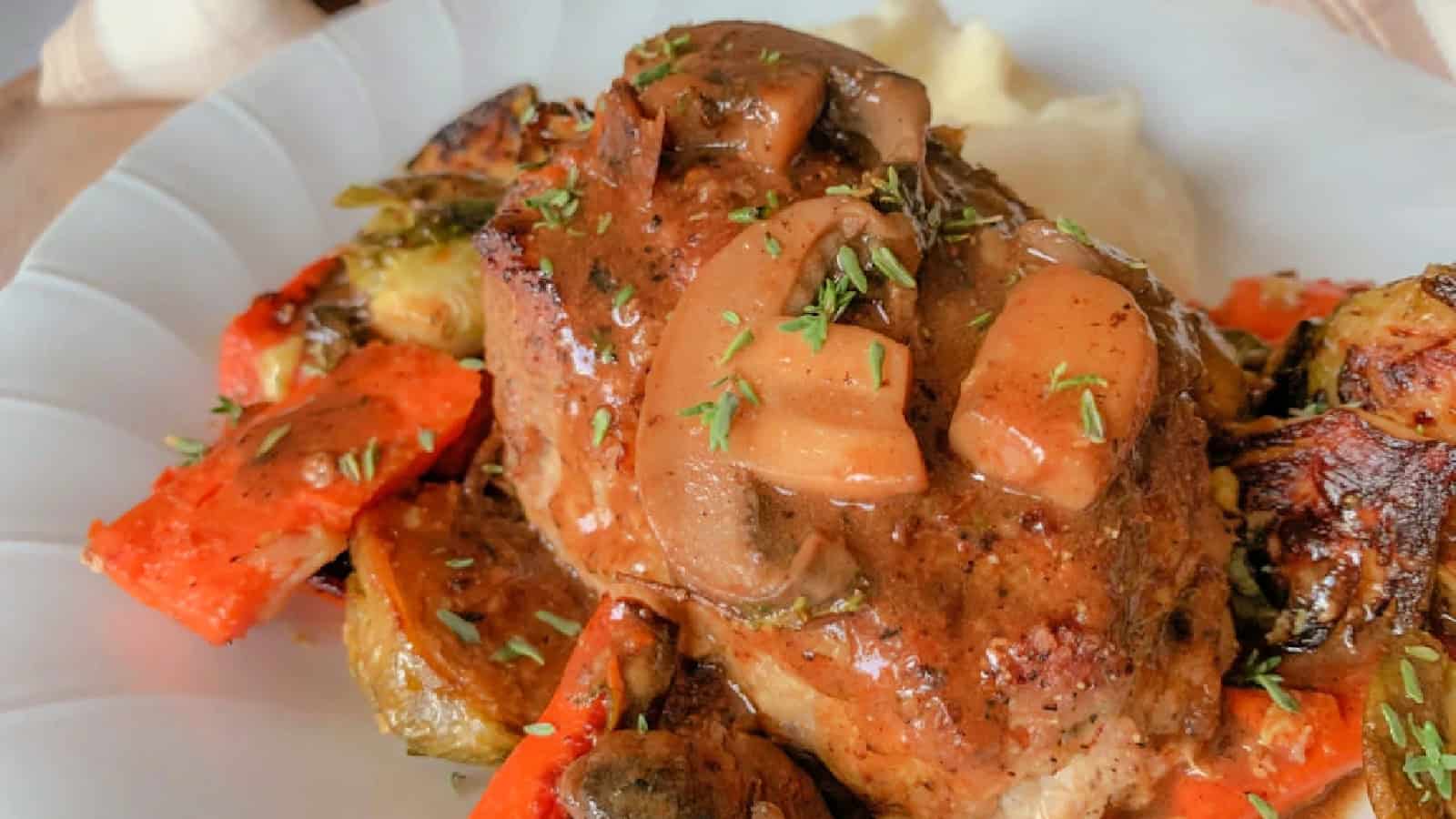 A savory dish featuring roasted pork topped with sliced mushrooms and herbs. It is served with roasted carrots and Brussels sprouts on a white plate. In the background, there's a glimpse of mashed potatoes.