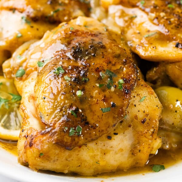 Close-up of a dish featuring seasoned chicken pieces in a savory sauce, garnished with herbs. Slices of lemon and green olives are visible in the sauce surrounding the chicken.