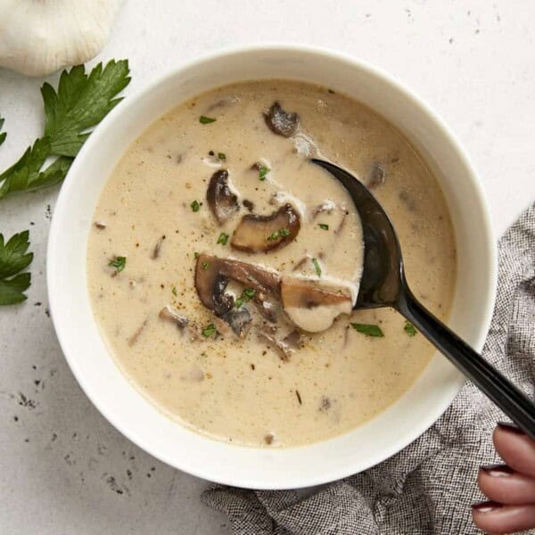 A bowl of creamy mushroom soup with sliced mushrooms and herbs, a black spoon, a hand holding the bowl, parsley, and garlic nearby.