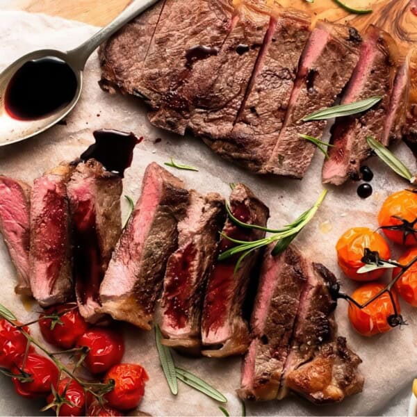 Sliced steak served on a wooden board, garnished with rosemary and cherry tomatoes, accompanied by a spoon filled with sauce. A small pile of French fries is visible on the side.