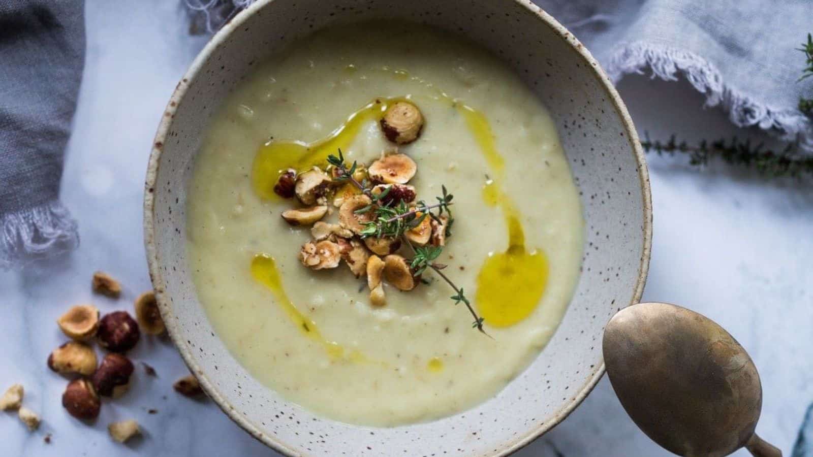 A bowl of creamy soup is topped with chopped hazelnuts, a drizzle of olive oil, and a sprig of thyme. A spoon rests on the marble surface next to the bowl, which is on a linen cloth.