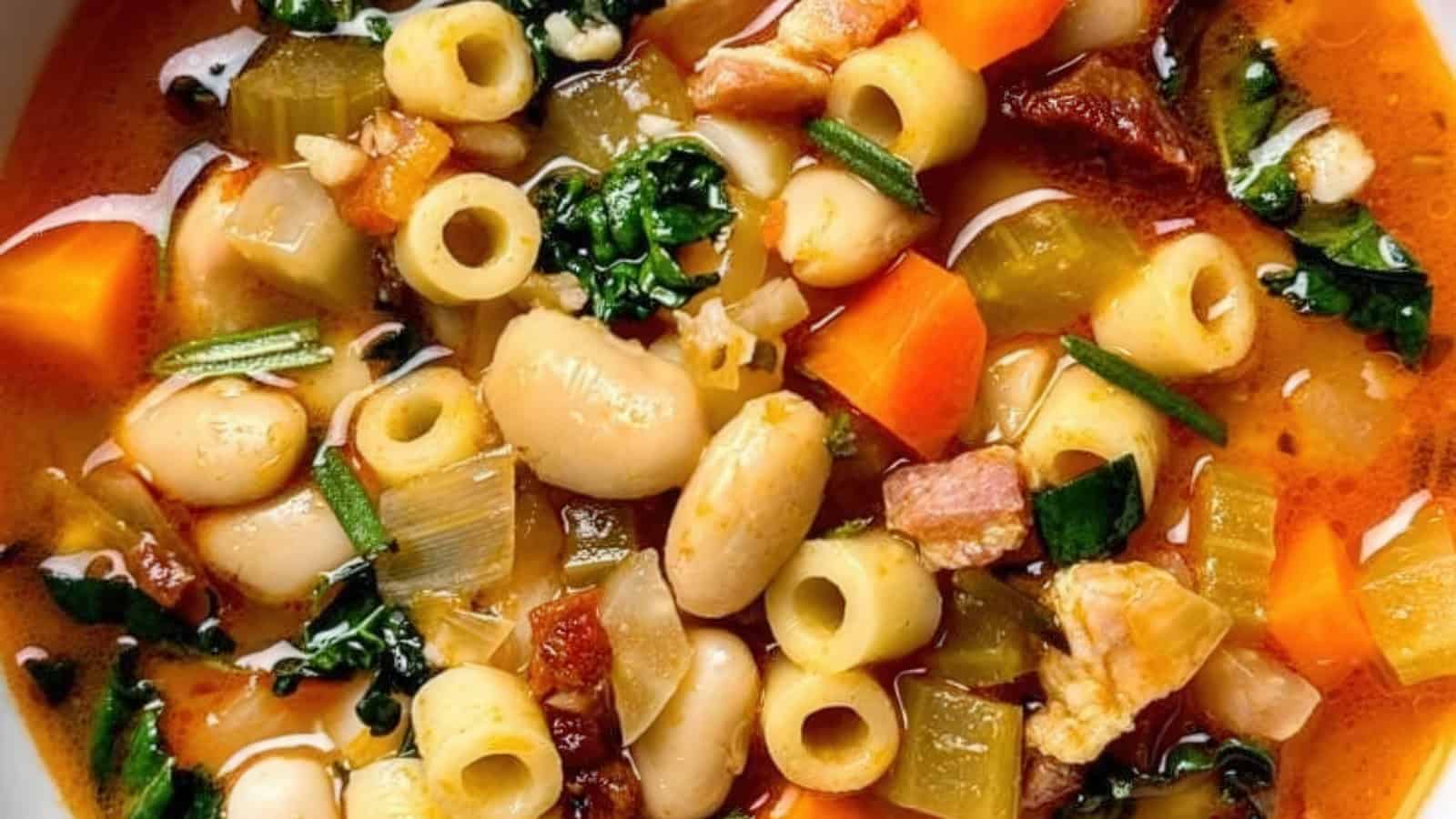 A close-up of a colorful soup featuring pasta, white beans, carrots, celery, greens, and bits of bacon in a rich, seasoned broth.