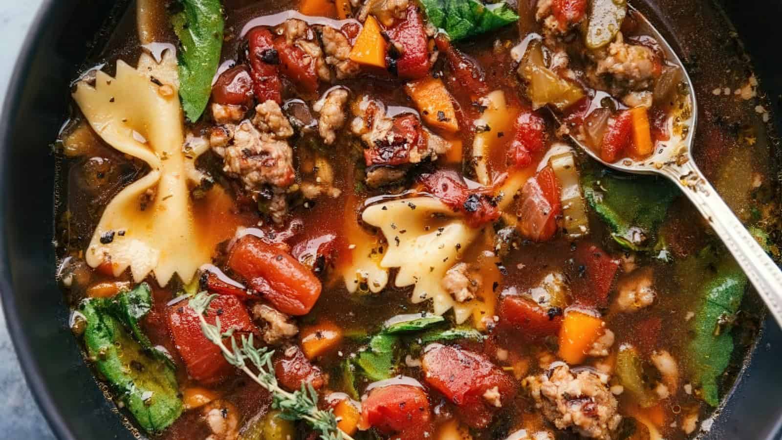 A hearty bowl of minestrone soup featuring bow tie pasta, ground beef, tomatoes, carrots, spinach, and herbs in a rich broth. A spoon rests in the soup, showcasing the colorful ingredients.