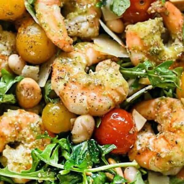 A vibrant salad featuring shrimp, arugula, cherry tomatoes, white beans, and shaved Parmesan cheese, all drizzled with a pesto dressing. The ingredients are mixed in a bowl, showcasing a fresh and colorful presentation.