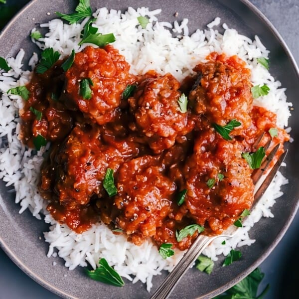 A plate of white rice topped with meatballs in a rich, red tomato sauce, garnished with fresh parsley. A fork rests on the plate. The background includes scattered parsley leaves.