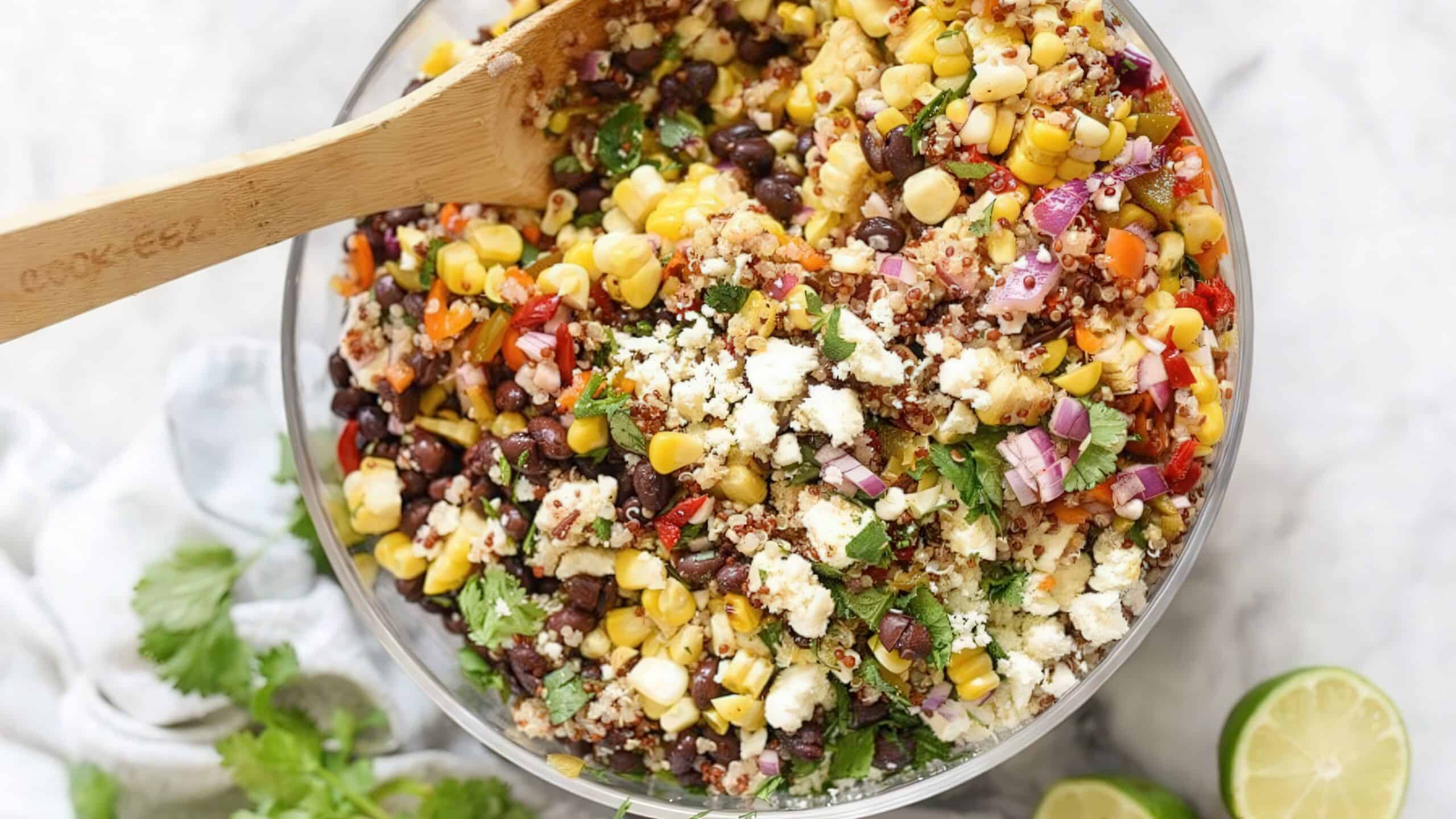 A bowl of colorful quinoa salad with black beans, corn, chopped red onions, diced red peppers, crumbled feta cheese, and cilantro. A wooden spoon rests in the bowl, with lime and cilantro on the side.
