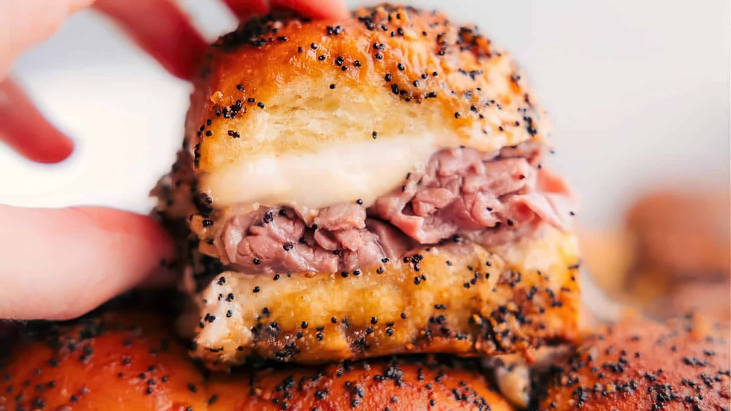 Close-up of a hand holding a sandwich made with a soft roll, stuffed with layers of roast beef and melted cheese. The bun is topped with poppy seeds and glazed with a buttery sauce.
