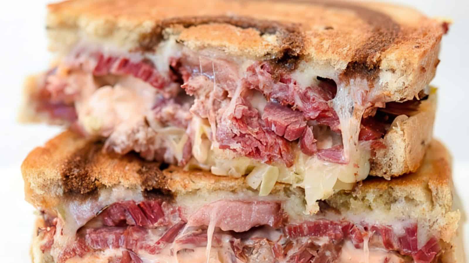 A close-up of a Reuben sandwich cut in half, stacked, and showing layers of corned beef, melted Swiss cheese, sauerkraut, and Russian dressing between slices of toasted rye bread.