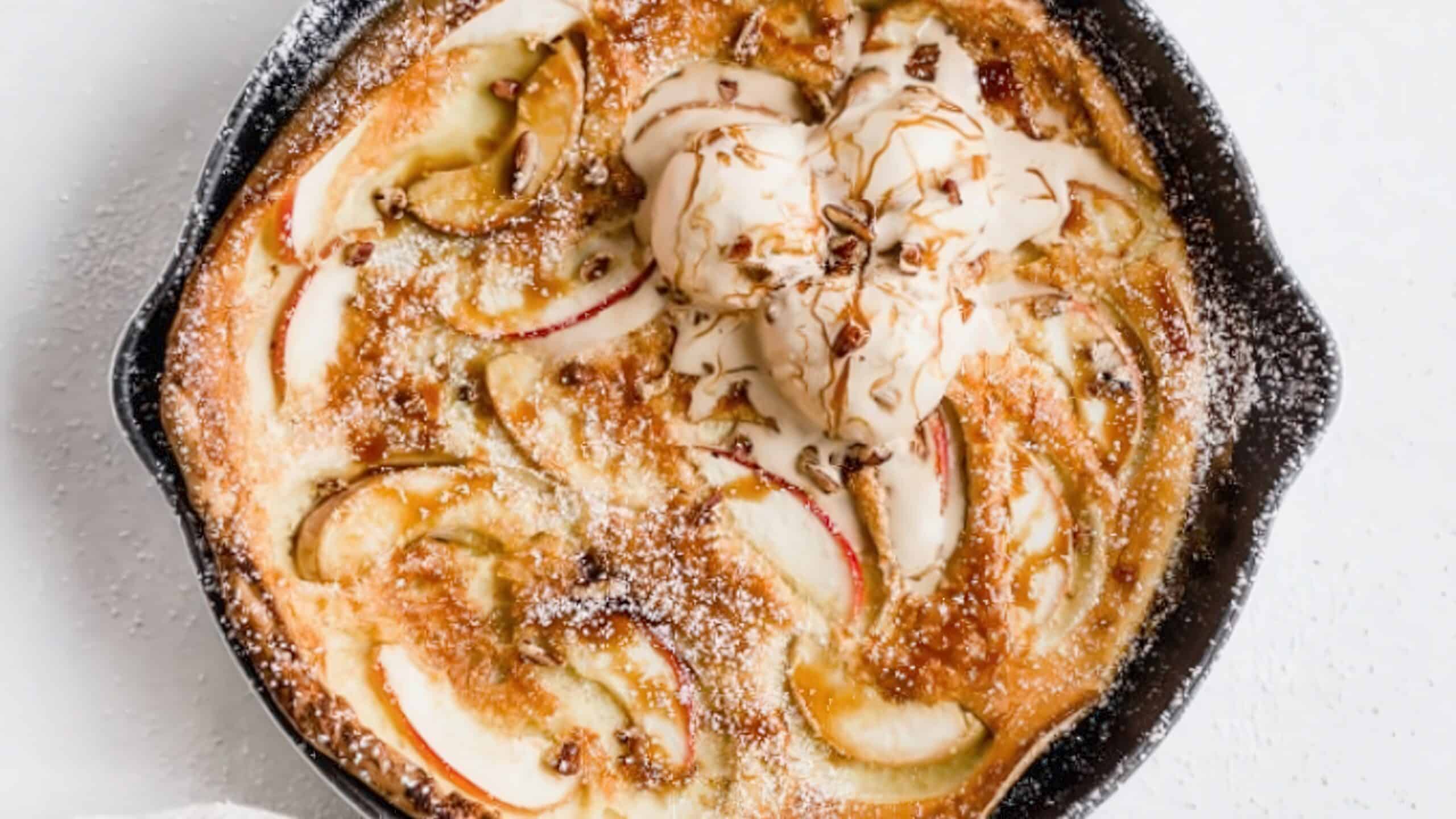 A skillet contains a baked apple dessert drizzled with caramel sauce, topped with a scoop of vanilla ice cream. The dish is dusted with powdered sugar and garnished with apple slices. It's set against a white background.