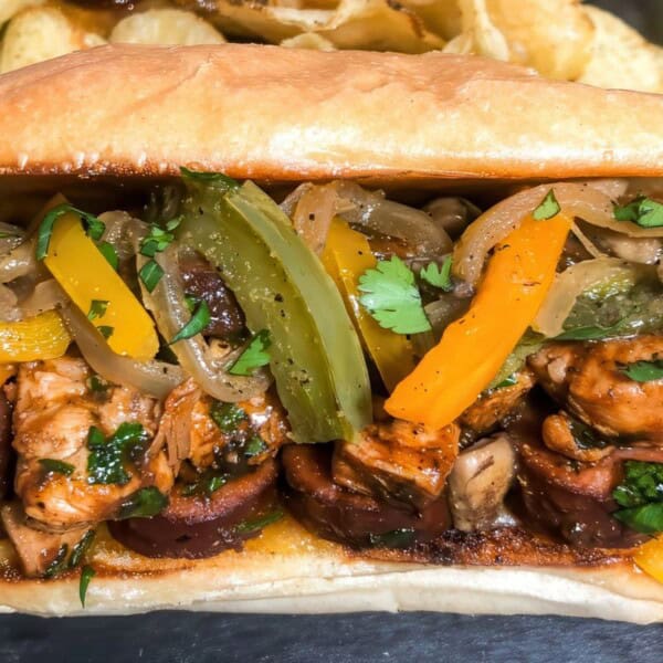 A toasted sandwich roll with grilled chicken, sausage, sautéed onions, bell peppers, fresh cilantro, and a side of potato chips.