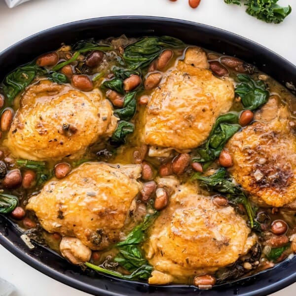 A black oval dish contains baked chicken thighs, spinach, and beans. Forks and a striped napkin are nearby.