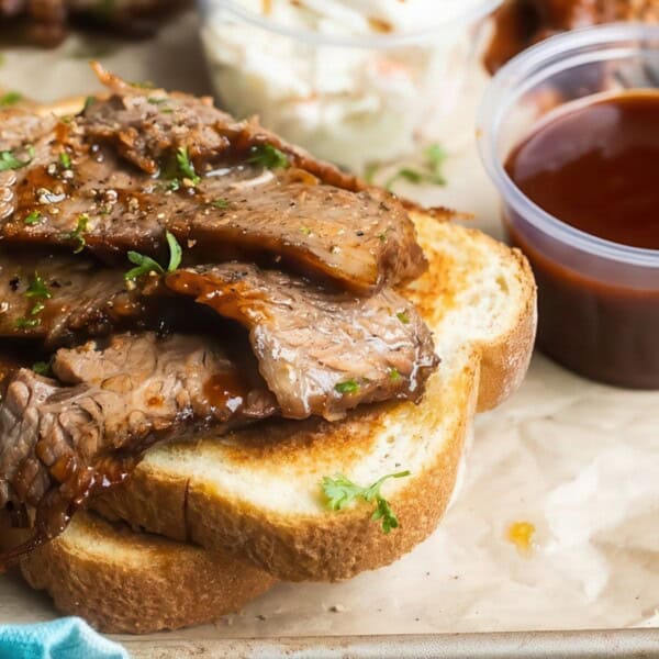 Slices of cooked brisket on white bread with herbs, barbecue sauce, and coleslaw in plastic containers on a tray.